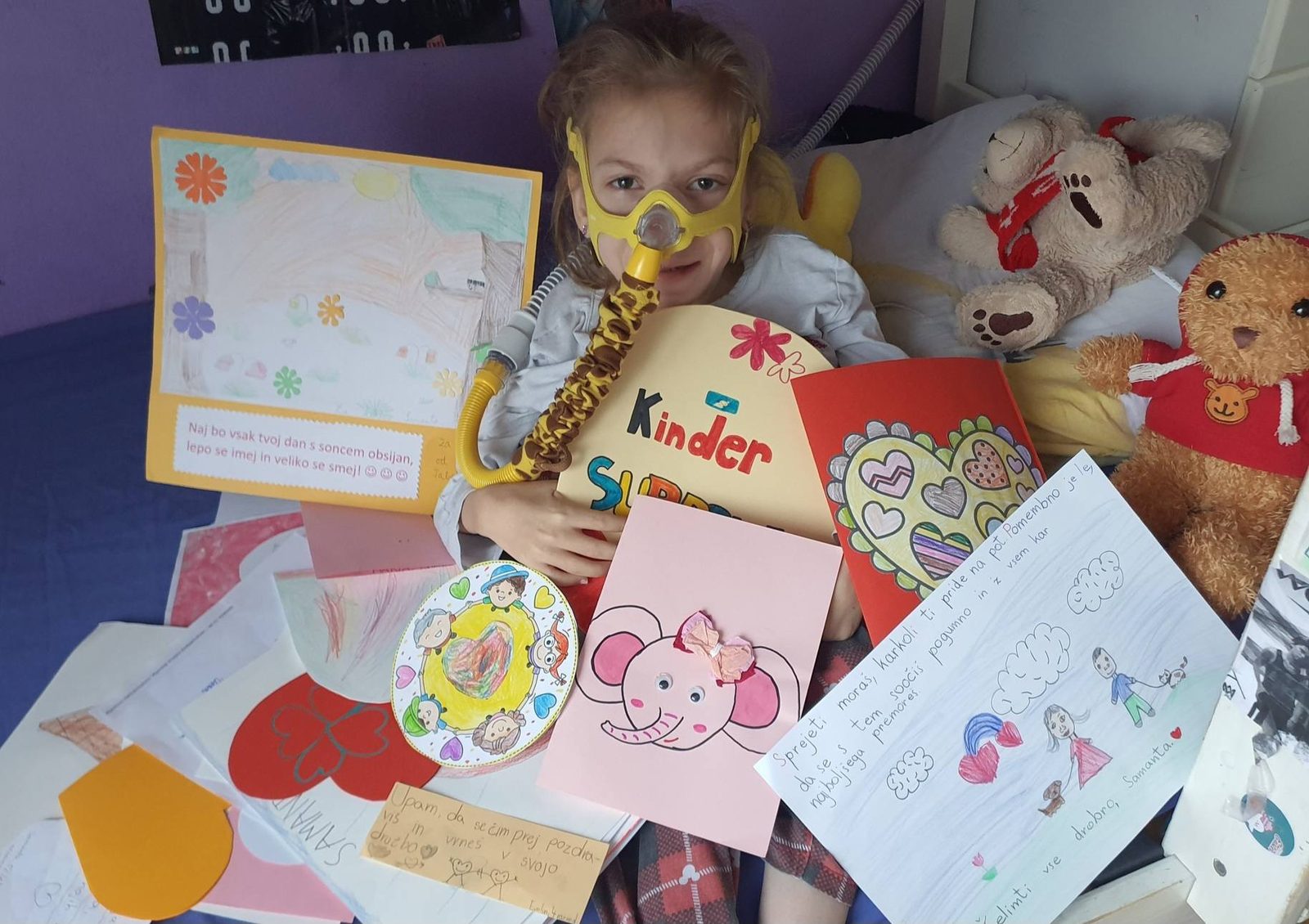 Deklica z dihalno masko med risbami in pismi, ki so ji jih poslali otroci iz vrtcev in šol